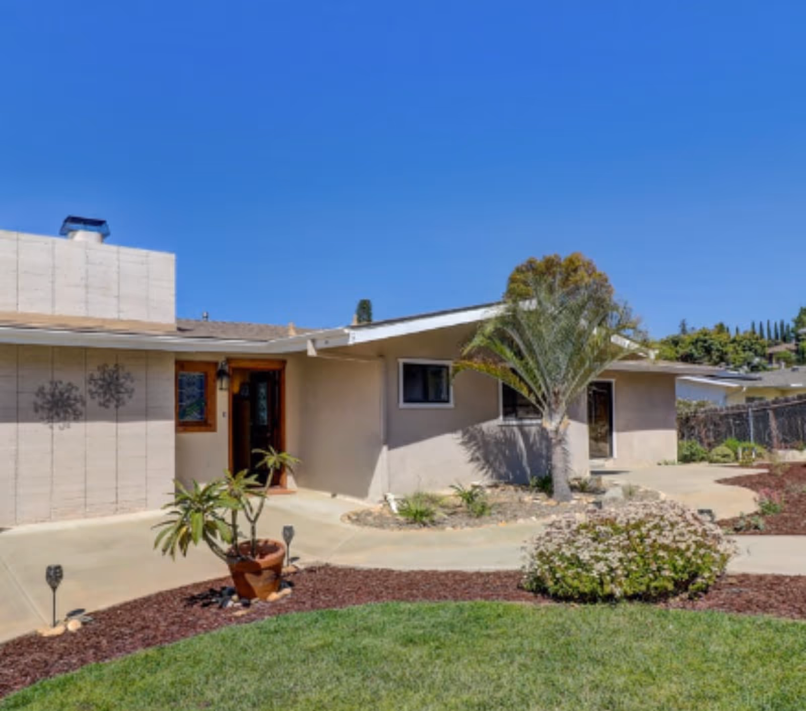 Exterior view of a single-story senior living facility building with a beige facade, a wooden door entrance, a small palm tree, potted plants, and landscaped garden beds with mulch and green grass under a clear blue sky.