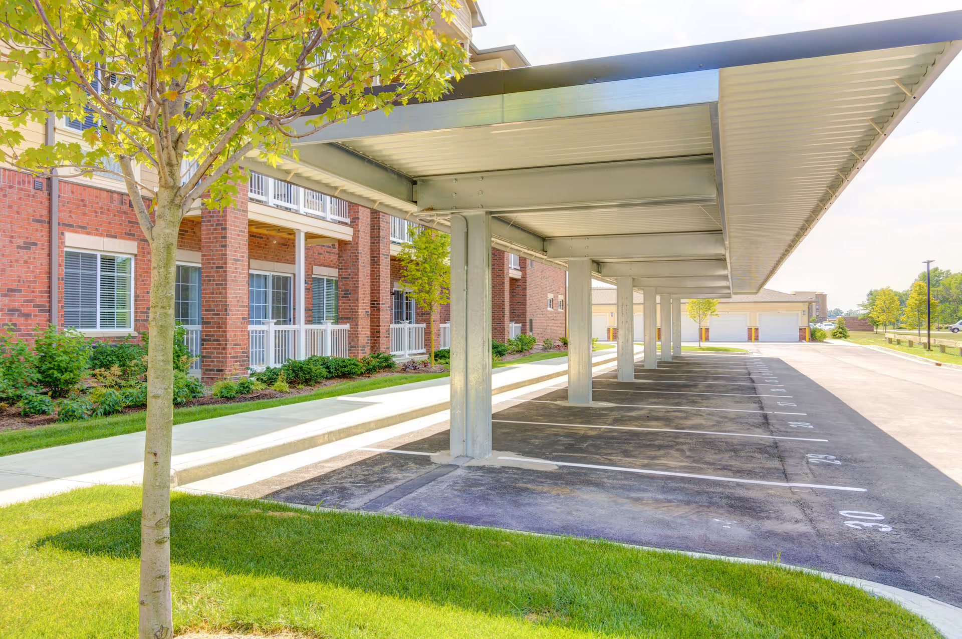Covered parking spaces next to a brick building with windows and small balconies, surrounded by green grass and trees under a clear sky.