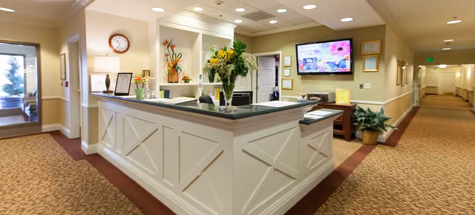 Reception desk area inside a senior living facility with a vase of flowers on the counter, a wall clock, a lamp, and a flat screen TV mounted on the wall. The hallway extends to the right with framed pictures on the wall and plants near the desk.