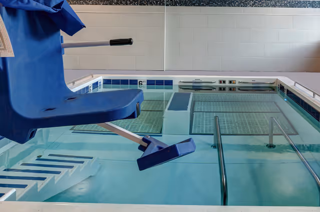Indoor therapeutic or exercise pool with a blue pool lift chair for accessibility, metal handrails, and steps leading into the water. The pool area has tiled walls and a clean, well-lit environment.