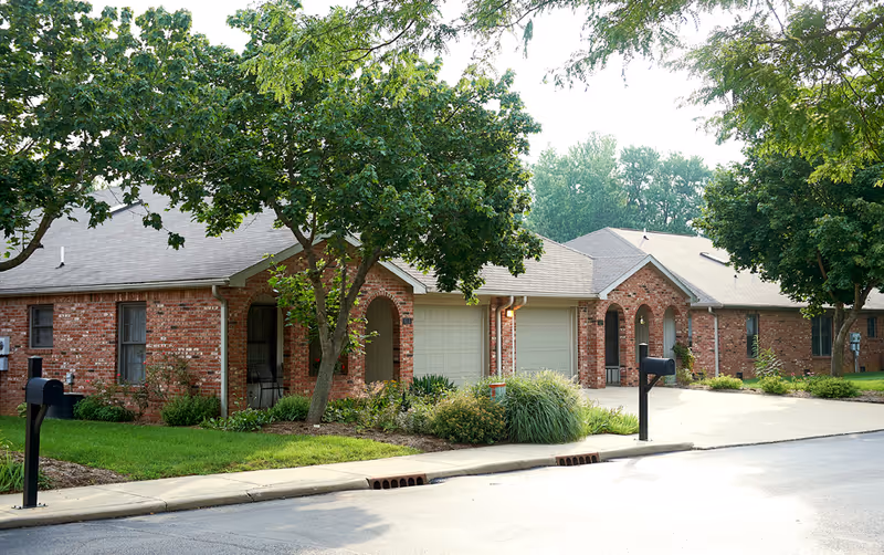 Exterior of a brick senior living facility with trees and arched front entrances.