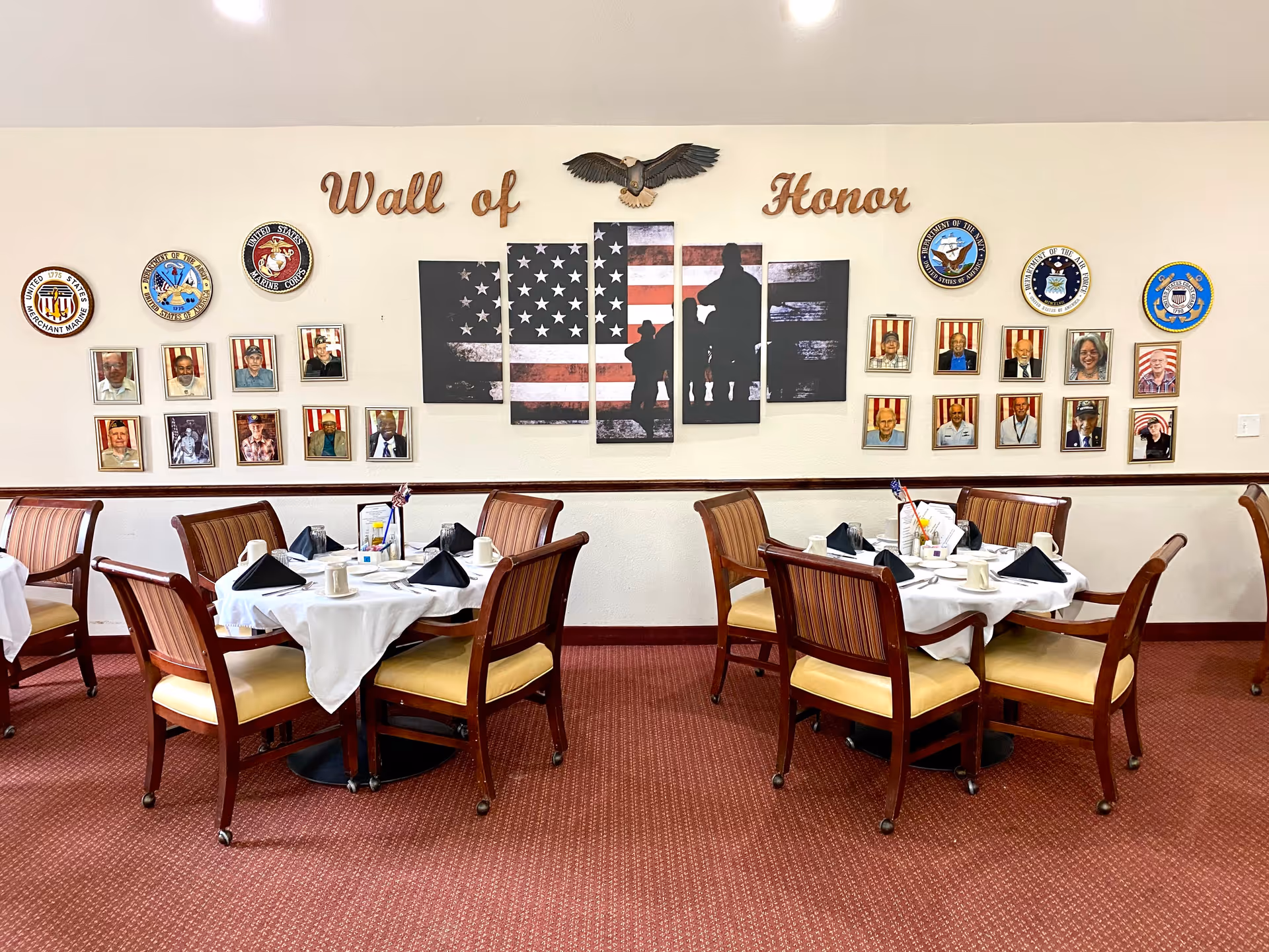 Dining room with two round tables set with white tablecloths, black napkins, cups, and condiments. Behind the tables is a wall display titled 'Wall of Honor' featuring an American flag artwork, an eagle decoration, military branch emblems, and framed photos of veterans.