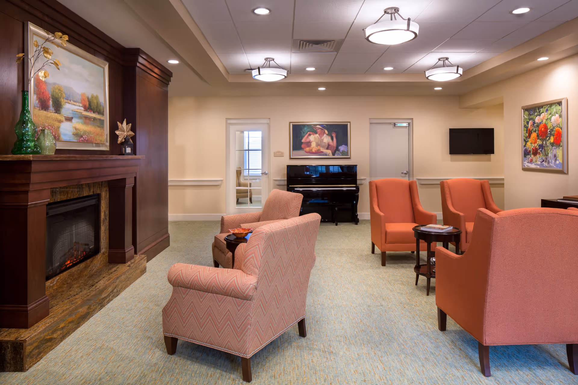 A cozy senior living facility lounge with four coral-colored armchairs arranged around a small round wooden table. There is a fireplace with a wooden mantle on the left side, decorated with a green vase, a small plant, and a framed landscape painting above it. The room has beige walls, a light blue carpet, and ceiling lights. At the back, there is a black upright piano beneath a framed painting of a woman, two doors, and a wall-mounted TV on the right side.