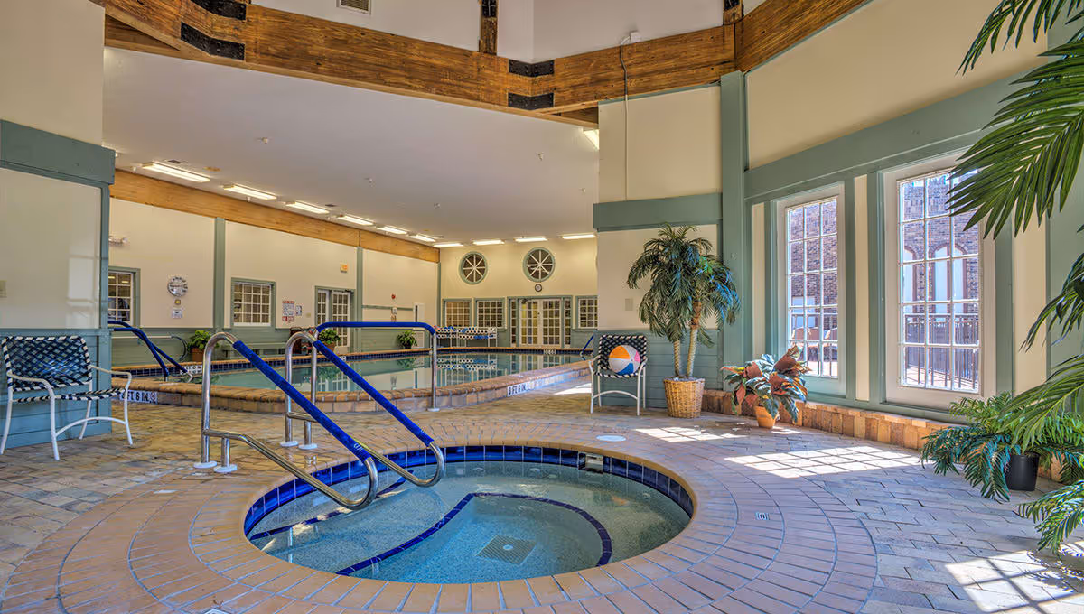 Indoor pool area with a hot tub in the foreground, surrounded by tiled flooring and several chairs. Large windows on the right side allow natural light to fill the space, and there are potted plants placed around the room. The pool has metal handrails and the walls are painted in light colors with wooden accents near the ceiling.
