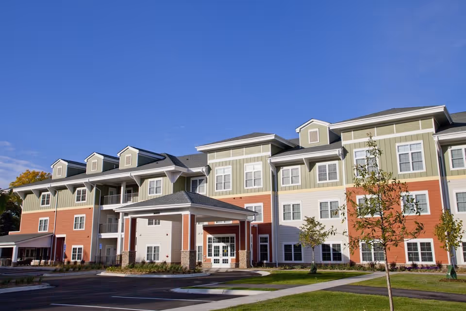 Exterior view of a multi-story senior living facility building with a covered entrance, multiple windows, and a landscaped area with young trees and grass under a clear blue sky.