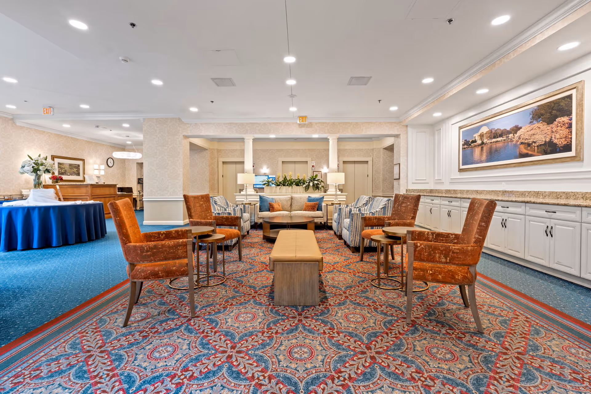 A spacious and well-lit lounge area in a senior living facility featuring a patterned carpet, several upholstered chairs in orange and striped blue, a beige sofa with colorful cushions, side tables, and lamps. The walls are decorated with framed artwork and floral arrangements, and there is a long countertop with cabinets underneath on the right side.