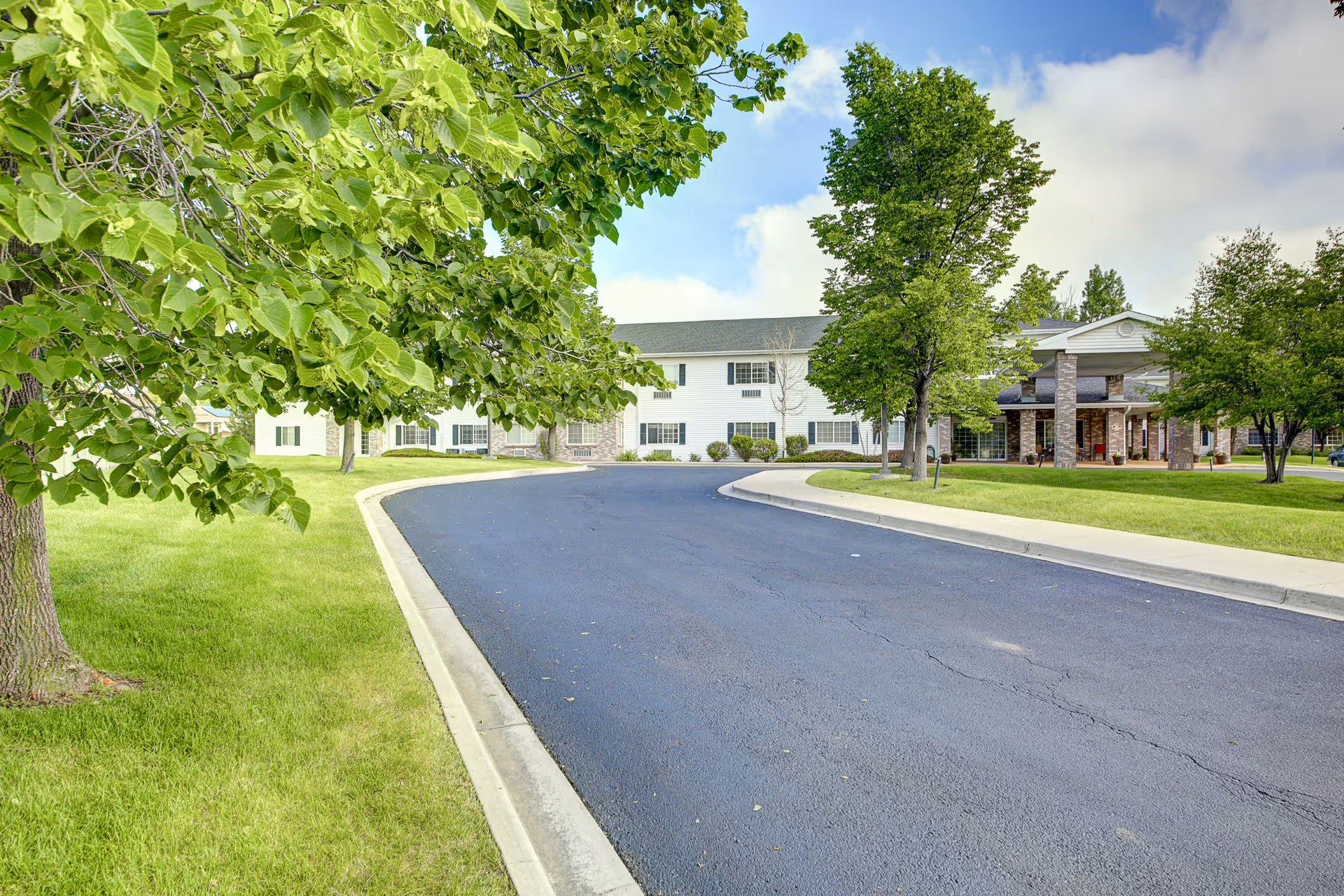 A paved driveway curves toward the front entrance of a two-story assisted living building surrounded by green lawns and trees.