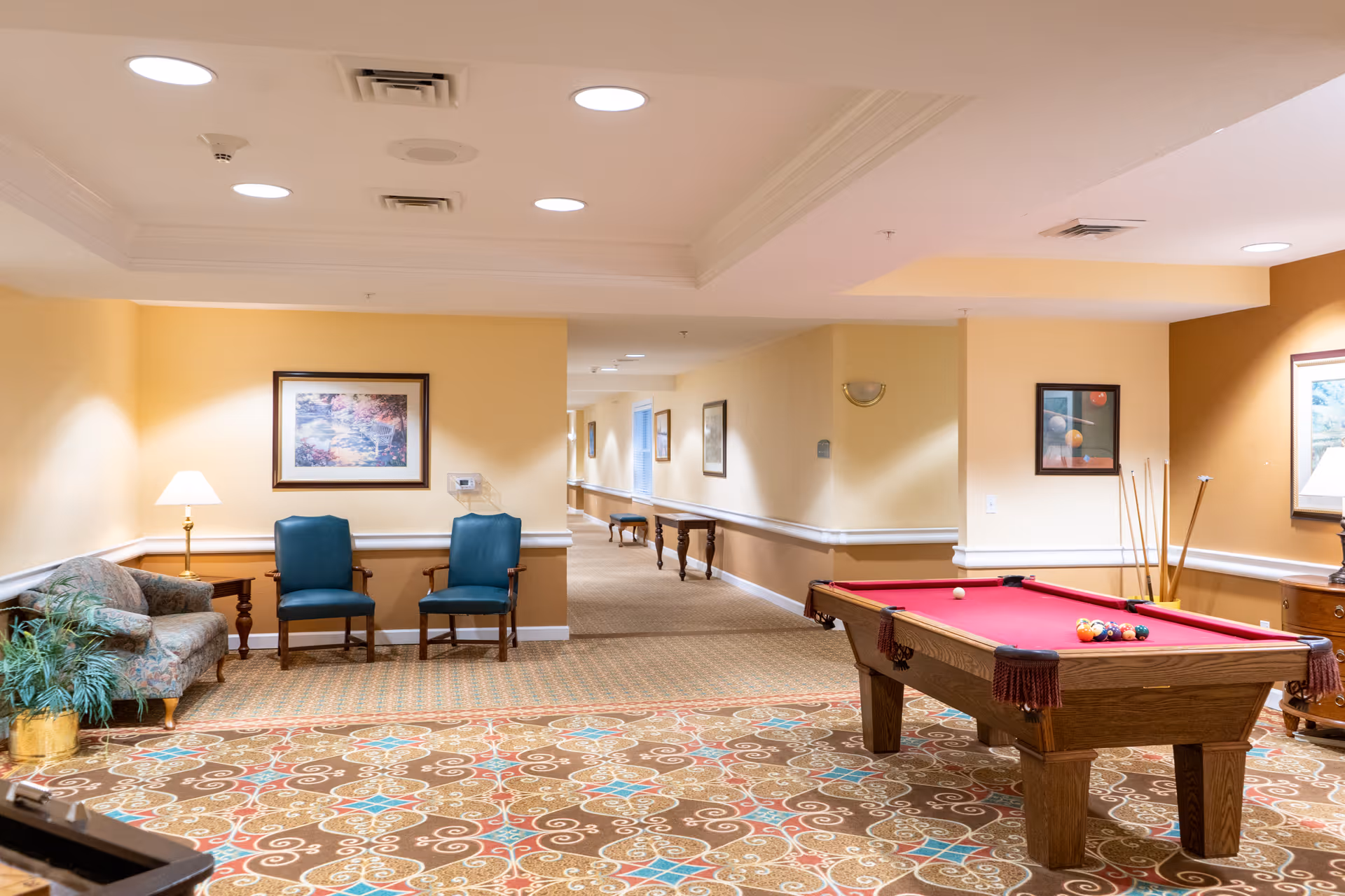 A cozy indoor common area with a red felt pool table, a patterned carpet, two blue chairs, a floral couch, a side table with a lamp, and framed artwork on the walls. The space is well-lit with ceiling lights and has a hallway extending into the background.