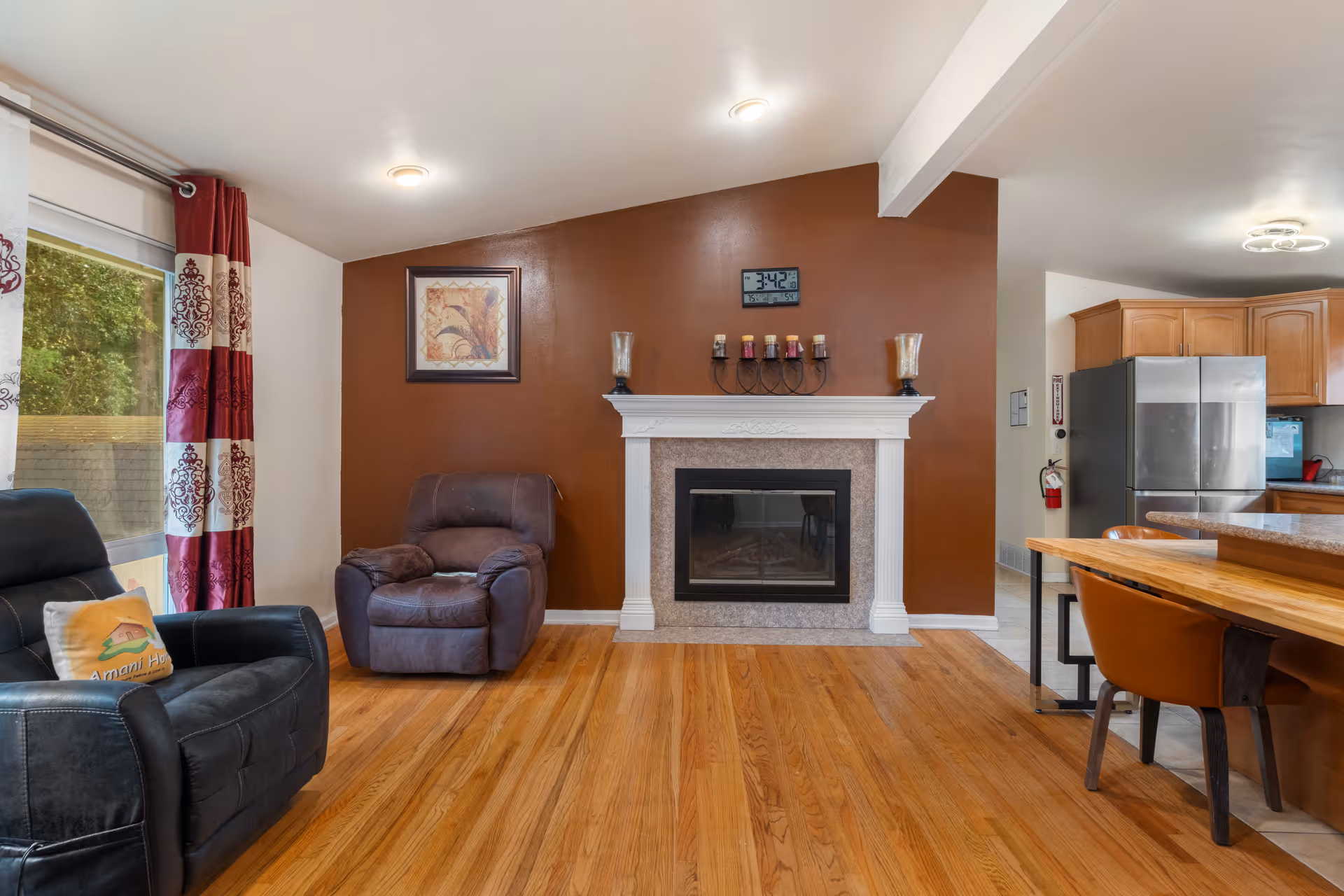 A cozy living room area with hardwood floors, featuring a brown recliner and a black leather armchair with a pillow that has 'Amani Home' written on it. There is a white fireplace with a brown accent wall behind it, decorated with candles and a framed picture. To the right, there is a wooden dining table with brown chairs and a kitchen area with wooden cabinets and a stainless steel refrigerator.