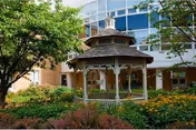 A wooden gazebo surrounded by lush green trees and colorful flowers in a garden area, with a multi-story building featuring large windows in the background.