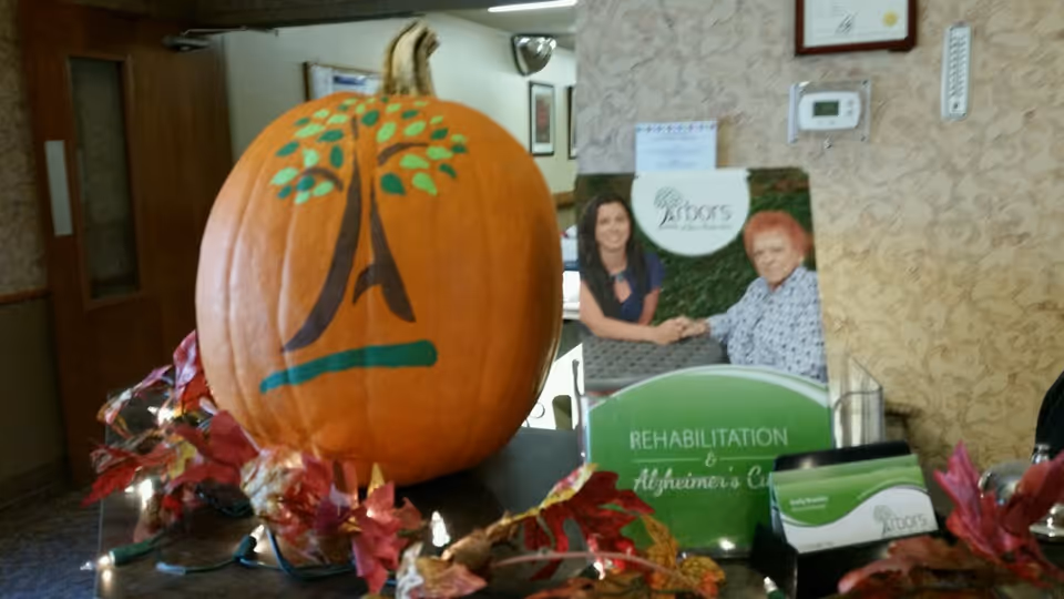 A large pumpkin painted with a tree logo sits on a table decorated with autumn leaves and string lights. Behind the pumpkin is a brochure holder displaying a flyer for Arbors at Stow, featuring two women holding hands and text about rehabilitation and Alzheimer's care. The setting appears to be an interior space with patterned wallpaper and a thermostat on the wall.