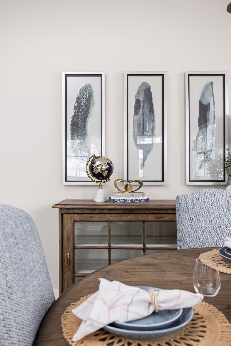 A dining area with a wooden table set with blue plates, cloth napkins, and a clear glass. Behind the table is a wooden cabinet with glass doors, topped with a decorative globe, a gold abstract sculpture, and stacked books. Three framed abstract art pieces hang on the light-colored wall above the cabinet.