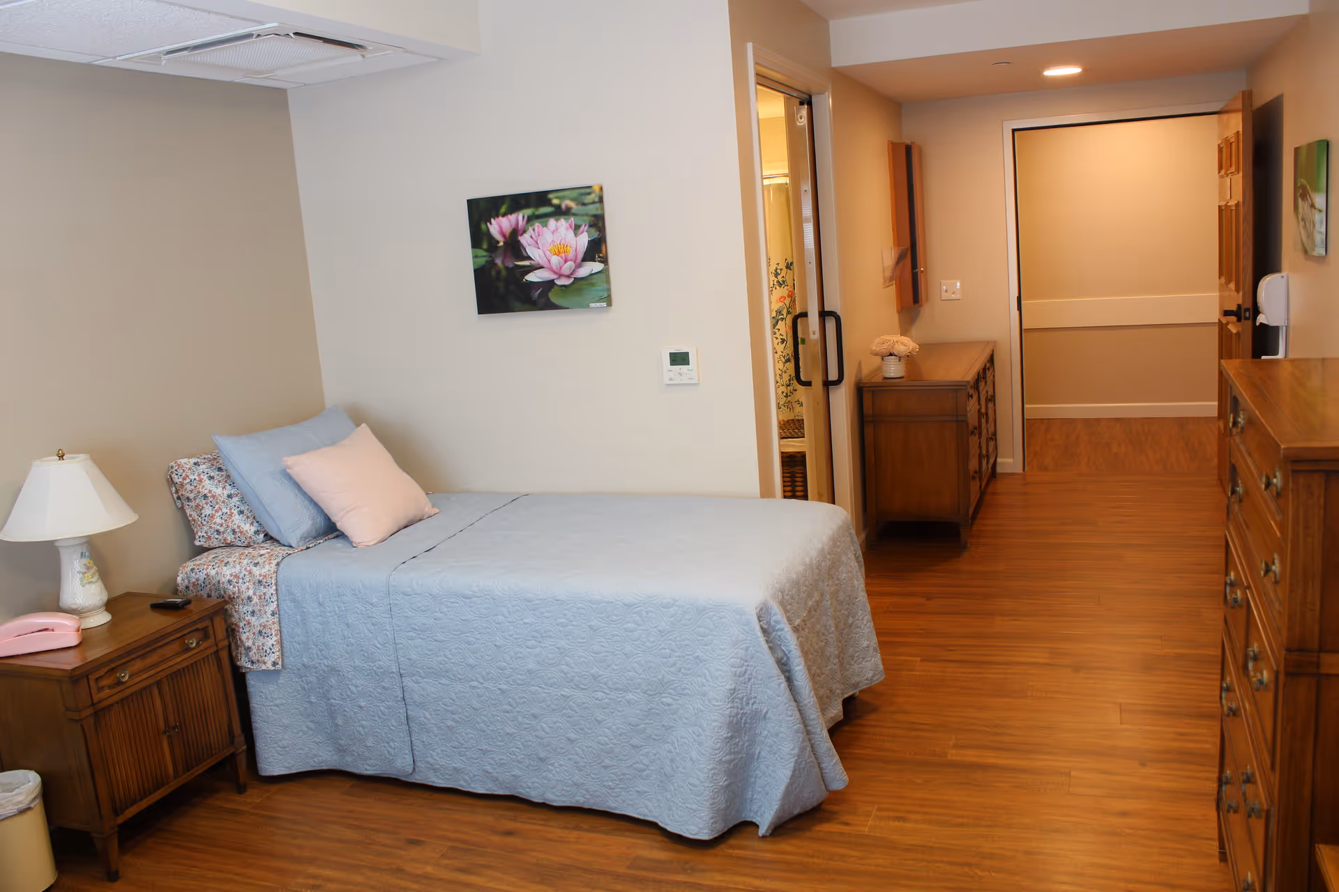 A neatly made single bed with blue bedding and pillows in a room with wooden furniture including a nightstand with a lamp and a dresser. There is a floral painting on the wall above the bed. The room has wooden flooring and an open doorway leading to a hallway and a bathroom.