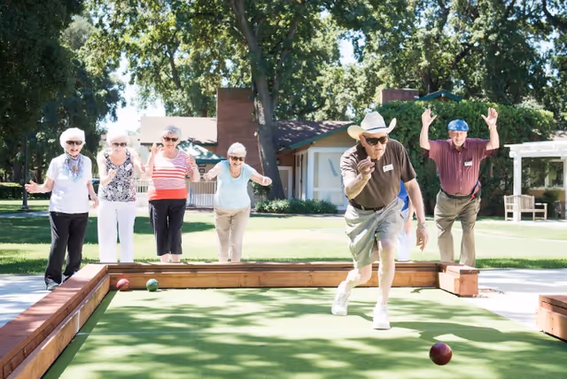 A group of older adults playing bocce on a green lawn court outside a senior living building.