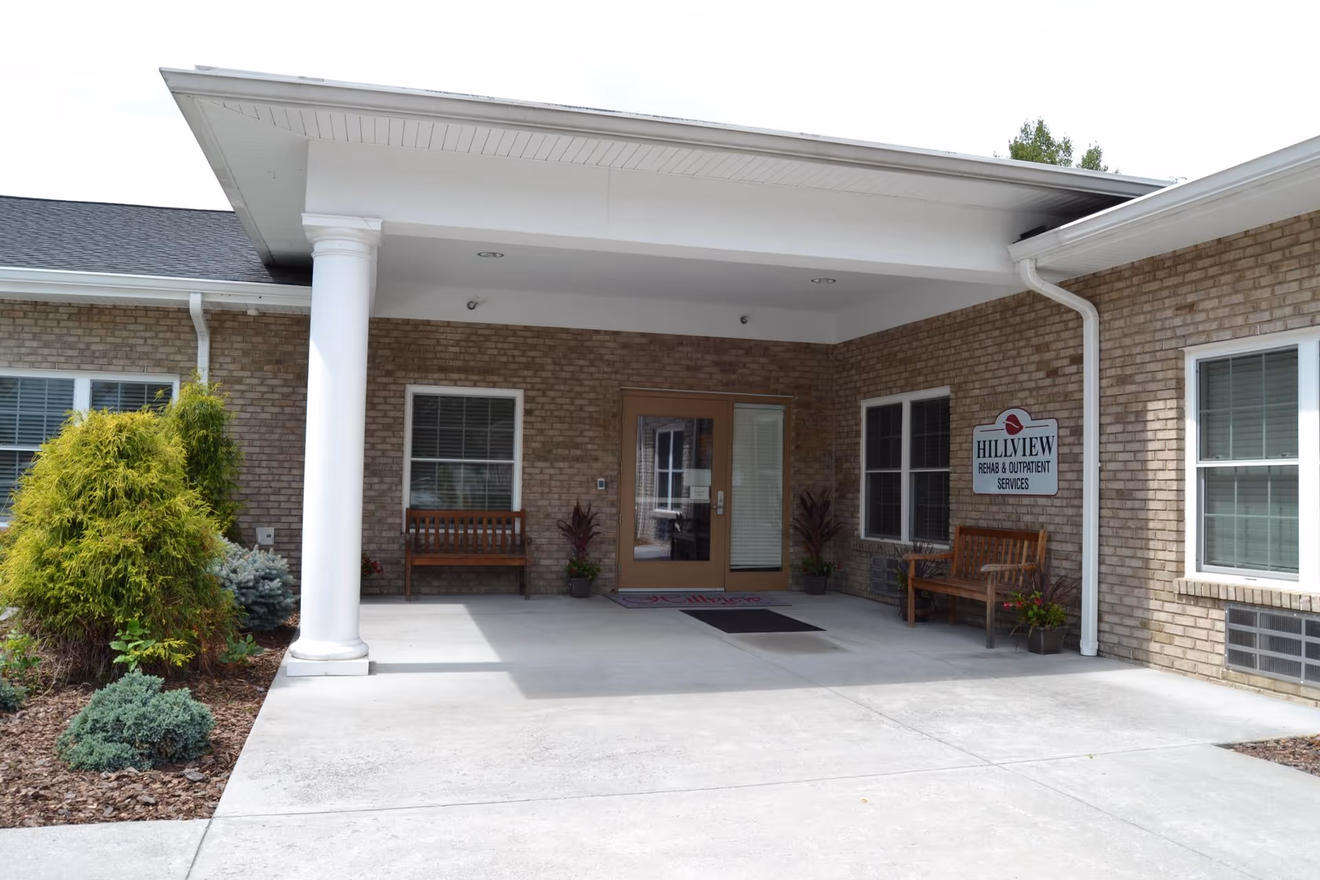 Entrance to Hillview Health and Rehabilitation Center showing a covered porch area with two wooden benches, potted plants, and a sign on the brick wall that reads 'Hillview Rehab & Outpatient Services'.