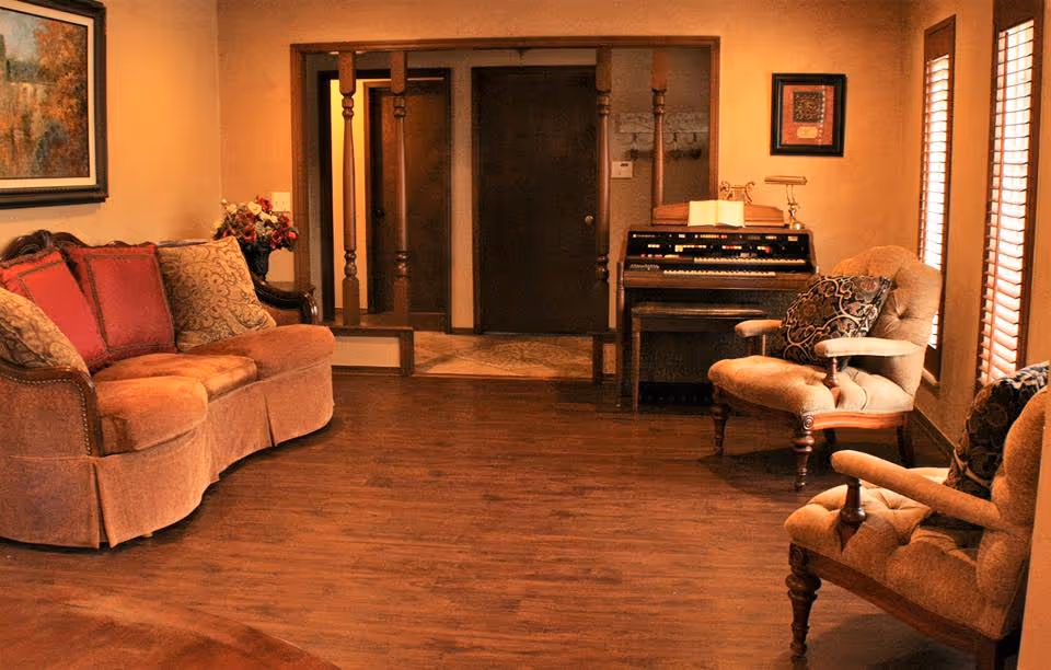 A cozy living room with a curved upholstered sofa, two armchairs, hardwood floors, and an organ against the back wall.
