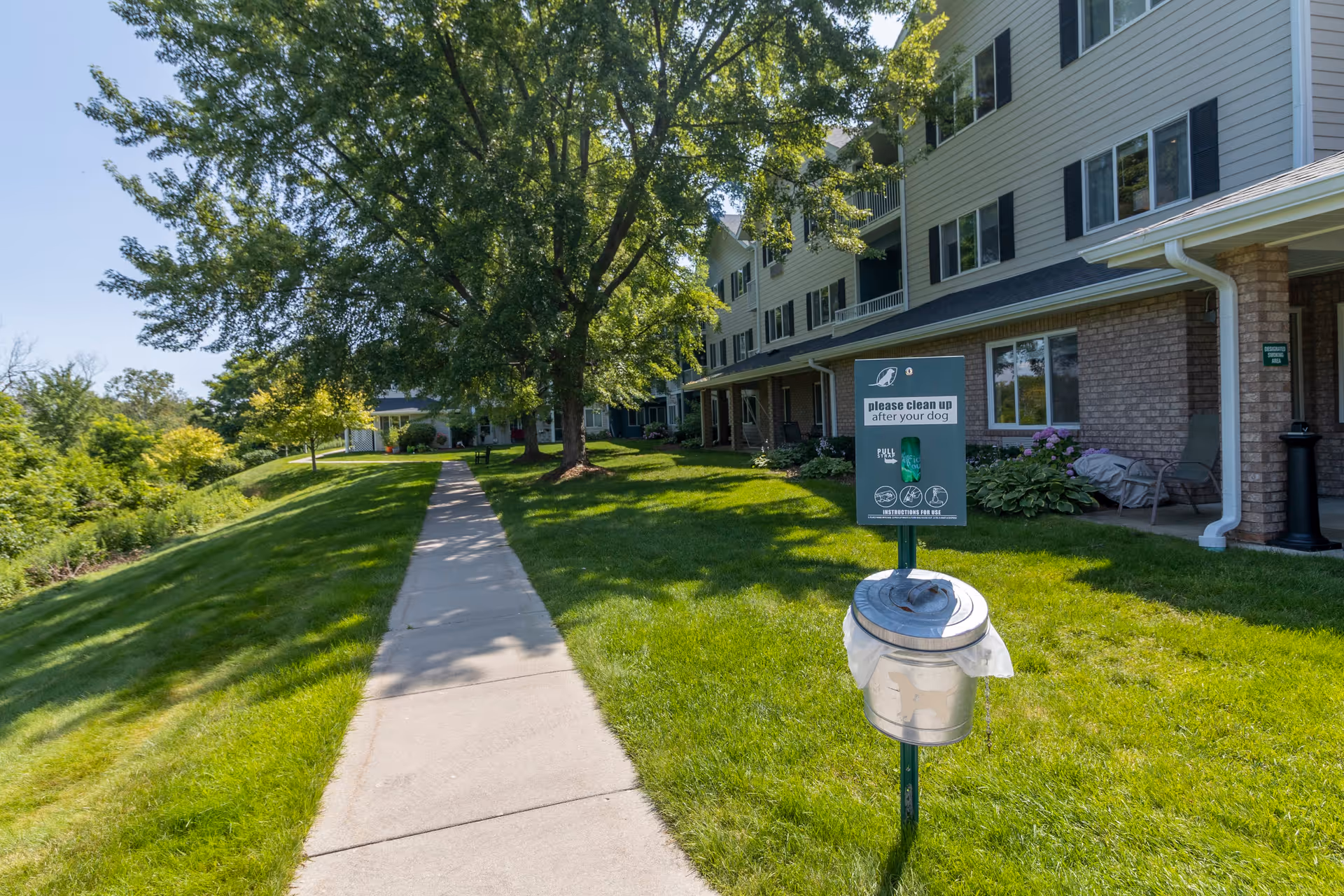 A sunny outdoor scene at Independence Village of Traverse City showing a paved walkway alongside a grassy area with trees and a multi-story residential building. A sign and container for dog waste bags is visible near the walkway, with a message asking to clean up after dogs.