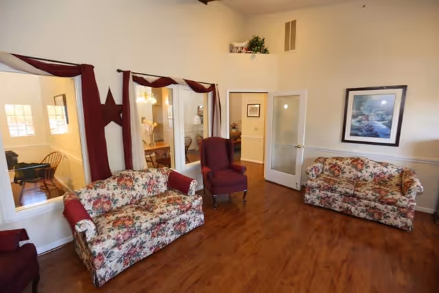 A cozy living room area with two floral-patterned sofas and two burgundy armchairs arranged on a wooden floor. The walls are light-colored with a framed landscape painting hanging on one side. There are windows with burgundy curtains and a glass door leading to another room.