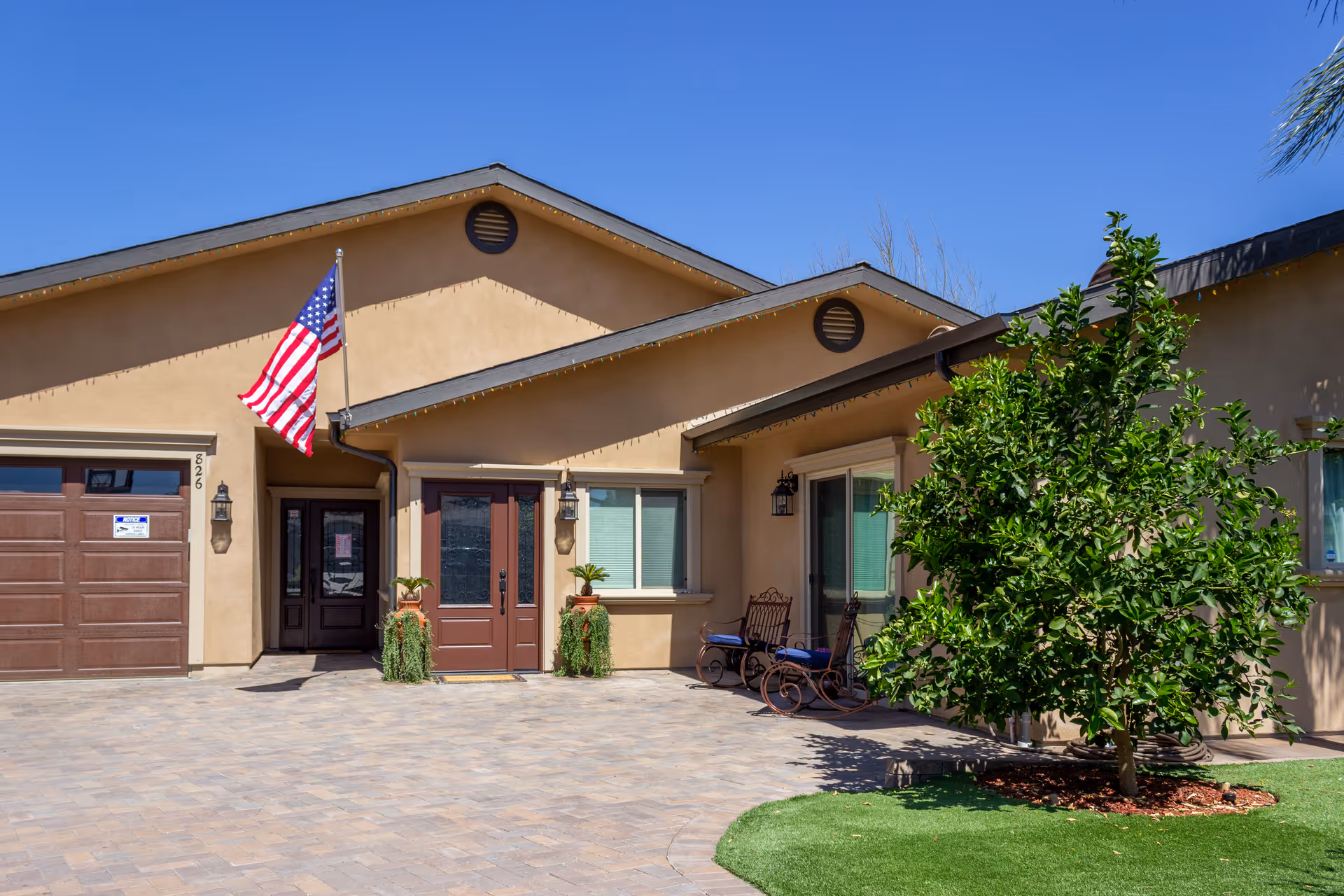 Exterior view of a single-story building with a beige facade, a brown garage door, and a main entrance with double doors. An American flag is mounted on a pole near the entrance. There are two potted plants flanking the front door and a small tree with green leaves on the right side near a seating area with two chairs and a small table. The ground is paved with stone tiles and there is a patch of green grass near the tree. The sky is clear and blue.