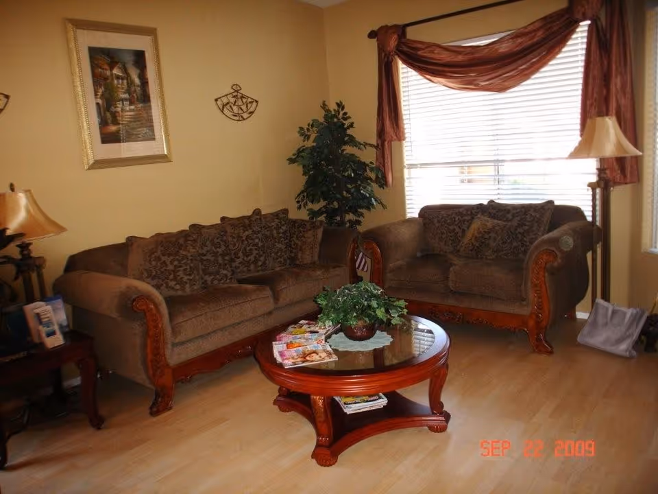 A cozy living room with two brown upholstered sofas featuring wooden carved armrests, a round wooden coffee table with a glass top and a potted plant centerpiece, a side table with a lamp and some items, a large window with blinds and sheer brown curtains, a floor lamp, a framed painting on the wall, and a decorative wall hanging.