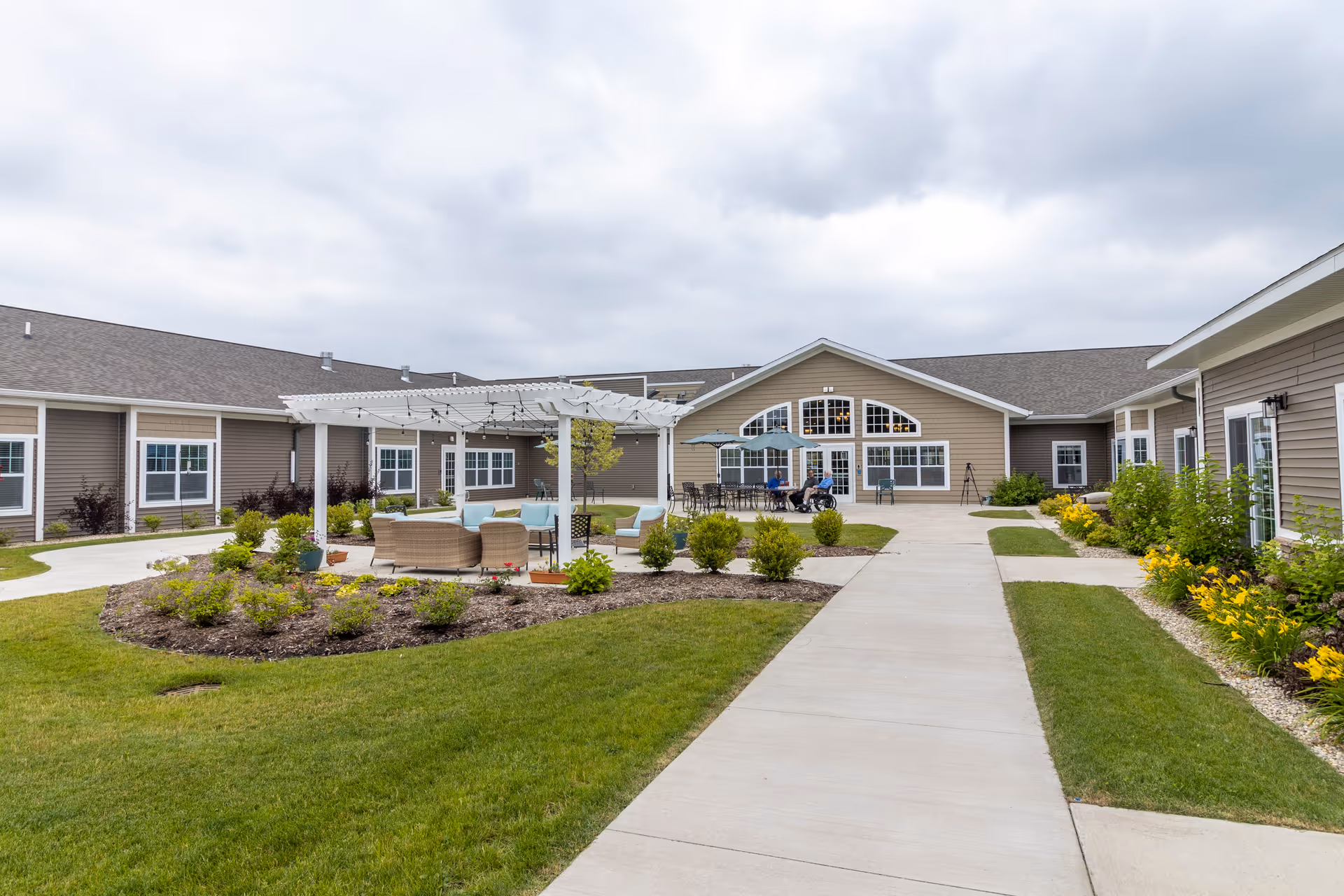 Courtyard of an assisted living facility with a pergola and outdoor seating, landscaped beds, and surrounding one-story buildings.