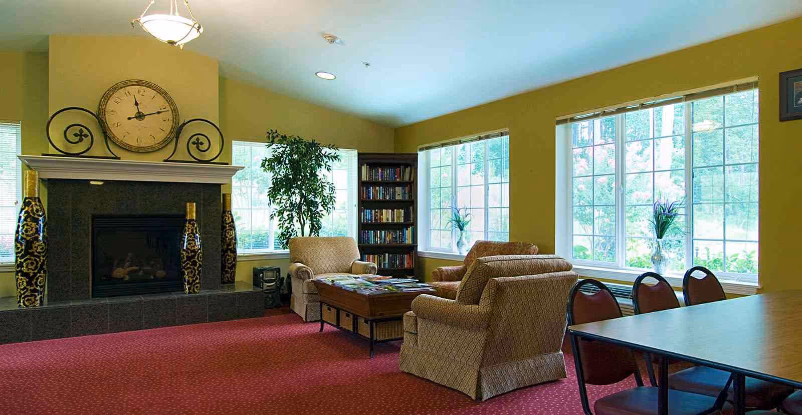 A cozy living room area with three upholstered armchairs arranged around a wooden coffee table covered with magazines. Behind the chairs is a fireplace with a large decorative clock above it and two tall ornamental vases on either side. Large windows with white frames let in natural light, and a bookshelf filled with books is positioned in the corner. A table with several chairs is visible on the right side of the room. The walls are painted yellow and the floor is covered with a red carpet.