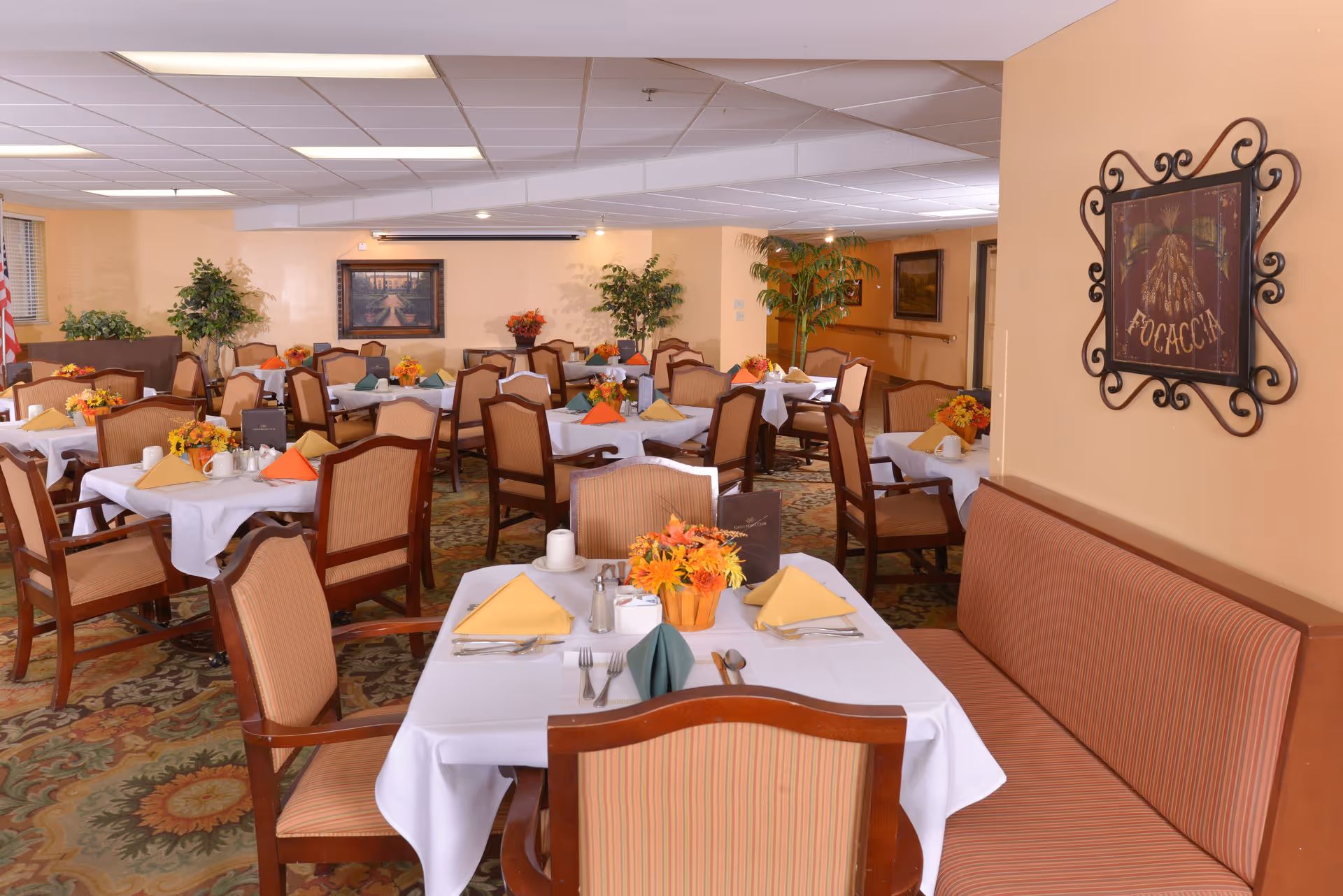 Dining room in a senior living facility with multiple tables covered in white tablecloths, each set with folded napkins in various colors, cutlery, and floral centerpieces. The room has cushioned chairs and a cushioned bench along one wall, with framed artwork and potted plants decorating the space.