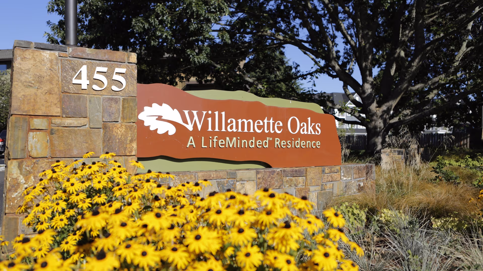 Stone sign for Willamette Oaks, a LifeMinded Residence, with the number 455 on the left side. The sign is surrounded by yellow flowers and greenery, with trees and a clear blue sky in the background.