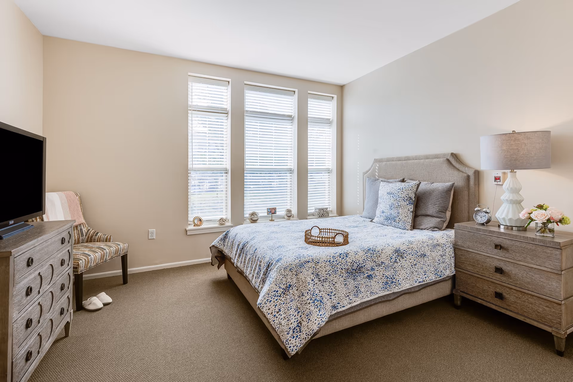 A bright bedroom with a large bed covered in a blue and white patterned bedspread, two gray pillows, and a decorative pillow. Next to the bed is a wooden nightstand with a white lamp, an alarm clock, and a small vase of flowers. Across from the bed is a wooden dresser with a flat-screen TV on top and a striped chair with white slippers on the floor beside it. Three tall windows with white blinds let in natural light.