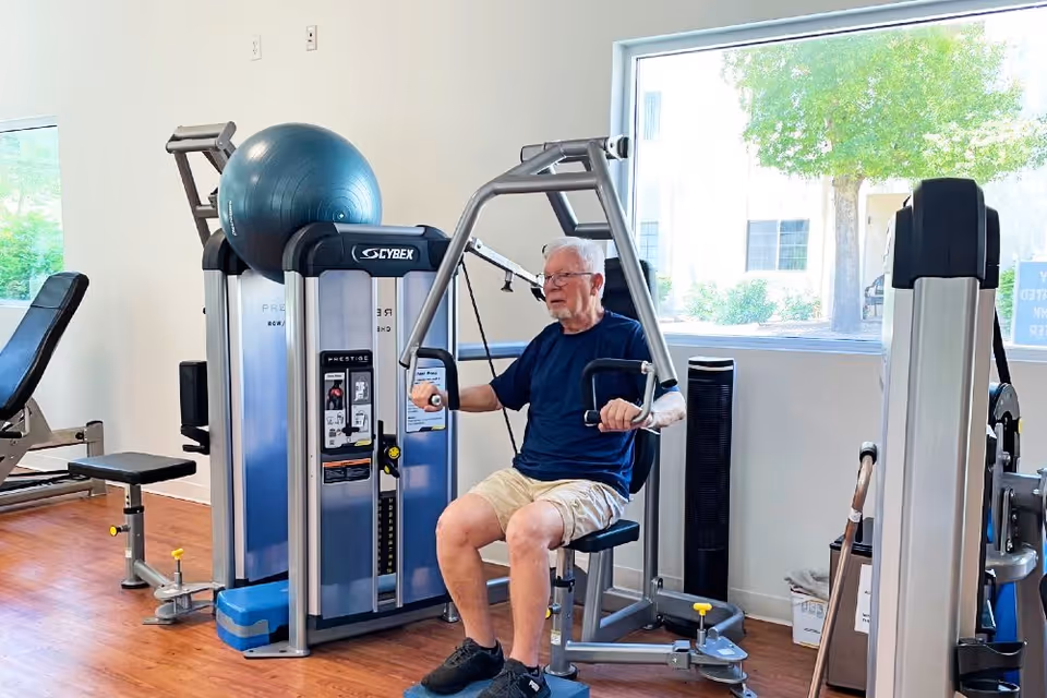 An older adult using a chest-press machine in a bright fitness room with large windows.