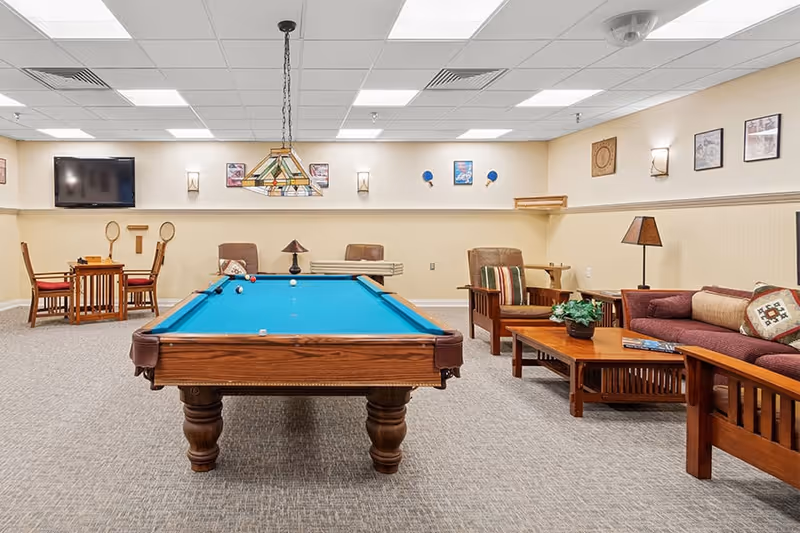 A recreational room with a pool table in the center, surrounded by wooden chairs and tables. There is a couch with cushions and a coffee table on the right side. The walls are decorated with framed pictures and a mounted TV is visible on the left. The room has a carpeted floor and a ceiling with fluorescent lights.