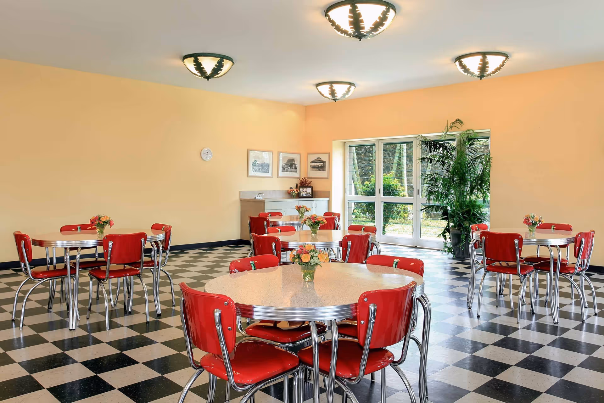 A bright dining room with checkered black and white flooring, round tables with chrome edges, and red chairs. Each table has a small vase with flowers. The walls are painted light yellow, and there are three ceiling lights. A large window and glass door let in natural light, with green plants visible outside. A small counter with framed pictures and a clock is on one wall.