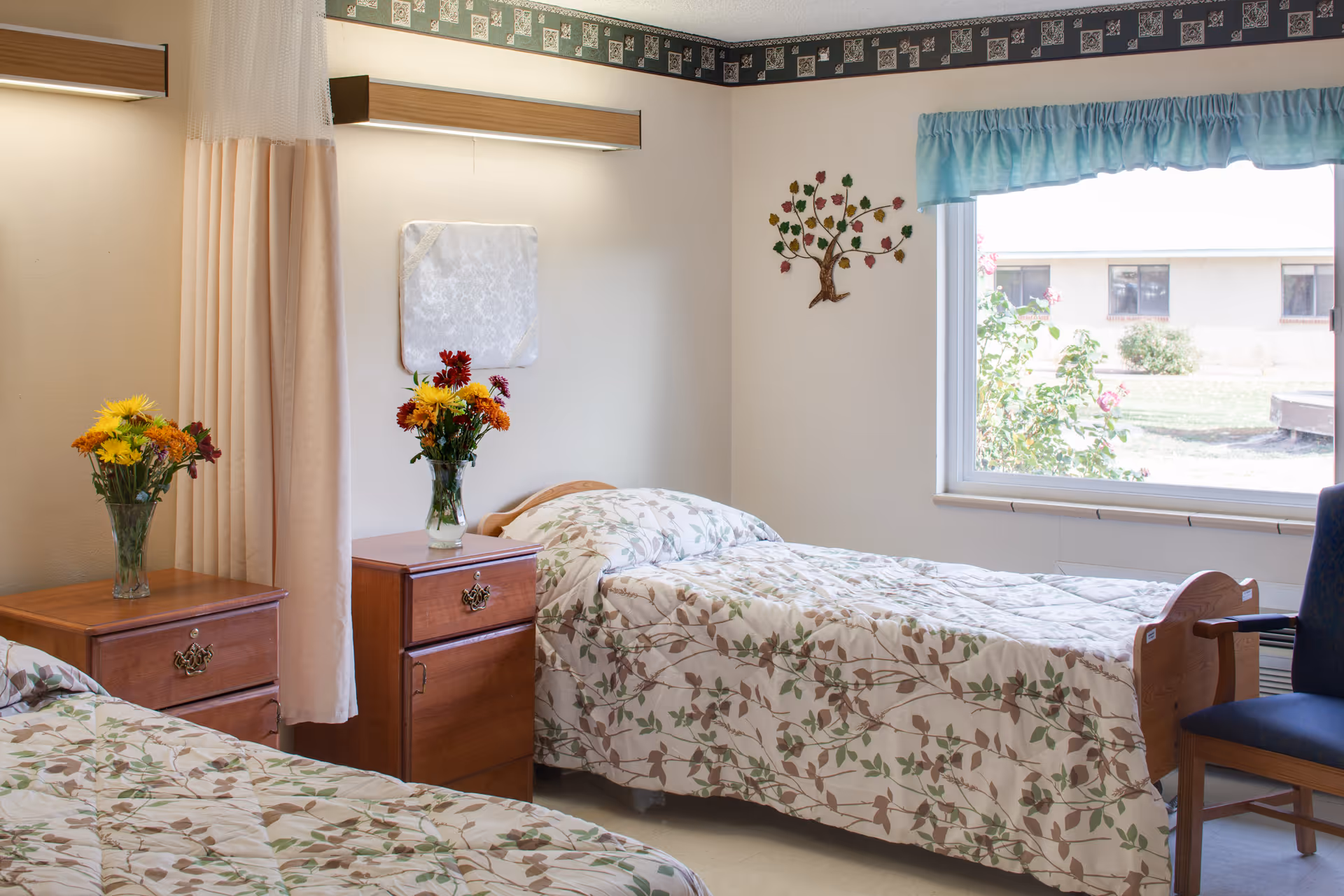 A bright and clean bedroom in a senior living facility with two single beds covered in floral patterned bedspreads. Each bed has a wooden nightstand beside it with a vase of colorful flowers. There is a large window with a blue valance letting in natural light, a blue chair near the window, and a decorative tree wall art on the wall.