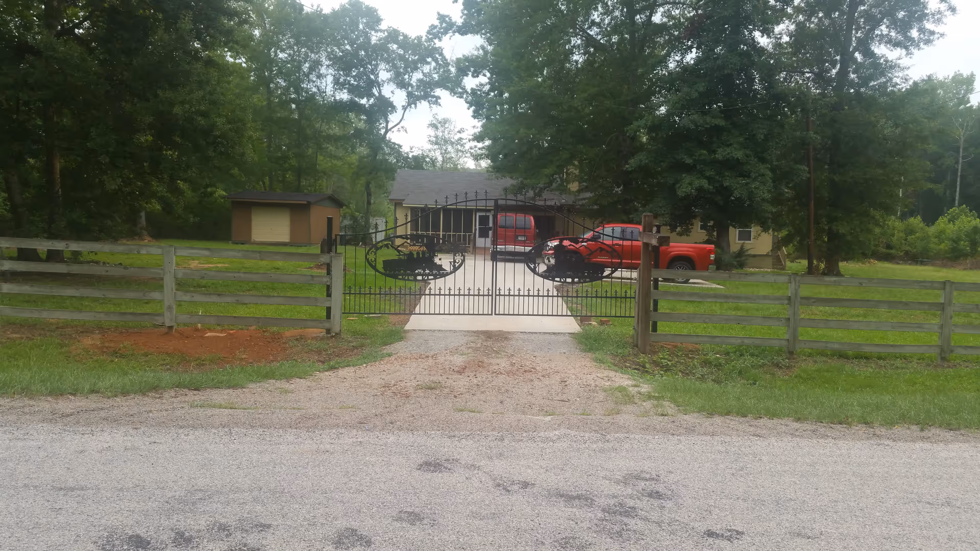 View of a gated driveway leading to a house with two red vehicles parked in front. The gate is black metal with decorative designs, and the property is surrounded by a wooden fence and trees.