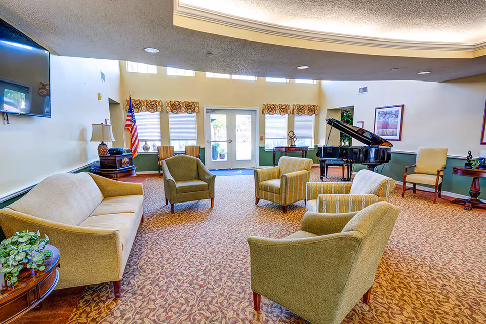 Bright communal lounge with sofas and armchairs arranged around a carpeted seating area and a grand piano near the entrance.