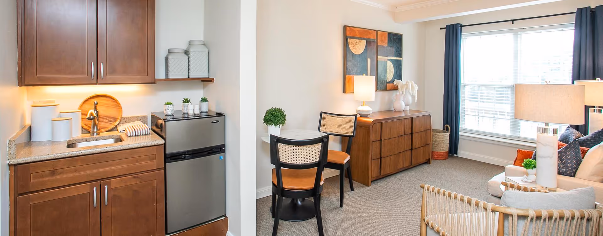 A cozy living area with a small kitchenette featuring wooden cabinets, a granite countertop, a sink, and a mini refrigerator. Adjacent to the kitchenette is a small round table with two chairs, a wooden sideboard with a lamp and decorative items, and a large window with dark curtains letting in natural light. The room is decorated with modern artwork and has a comfortable seating area with cushions and lamps.