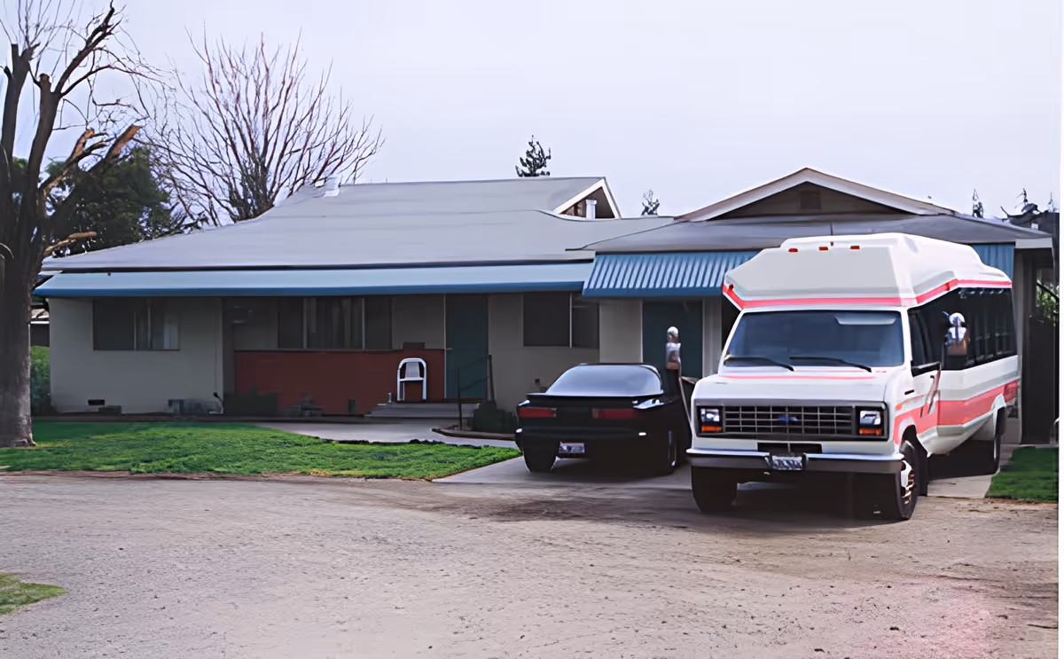 Exterior view of a single-story building with a gray roof and a blue awning over the entrance. There is a black car and a white and red shuttle bus parked in the driveway. A leafless tree is visible on the left side, and the ground is a mix of grass and dirt.