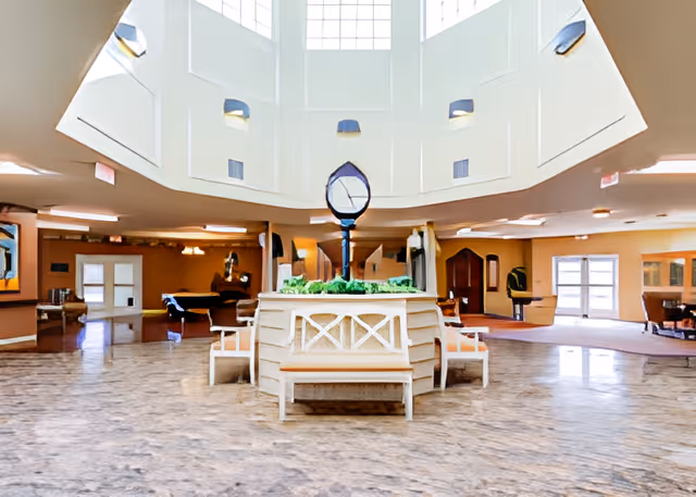 Spacious interior common area with a high ceiling and large windows allowing natural light. In the center, there is a decorative planter with greenery and a tall sculpture. Surrounding the planter are several wooden benches with white cushions. The floor is covered with a marble-like pattern, and there are multiple doorways and seating areas visible in the background.