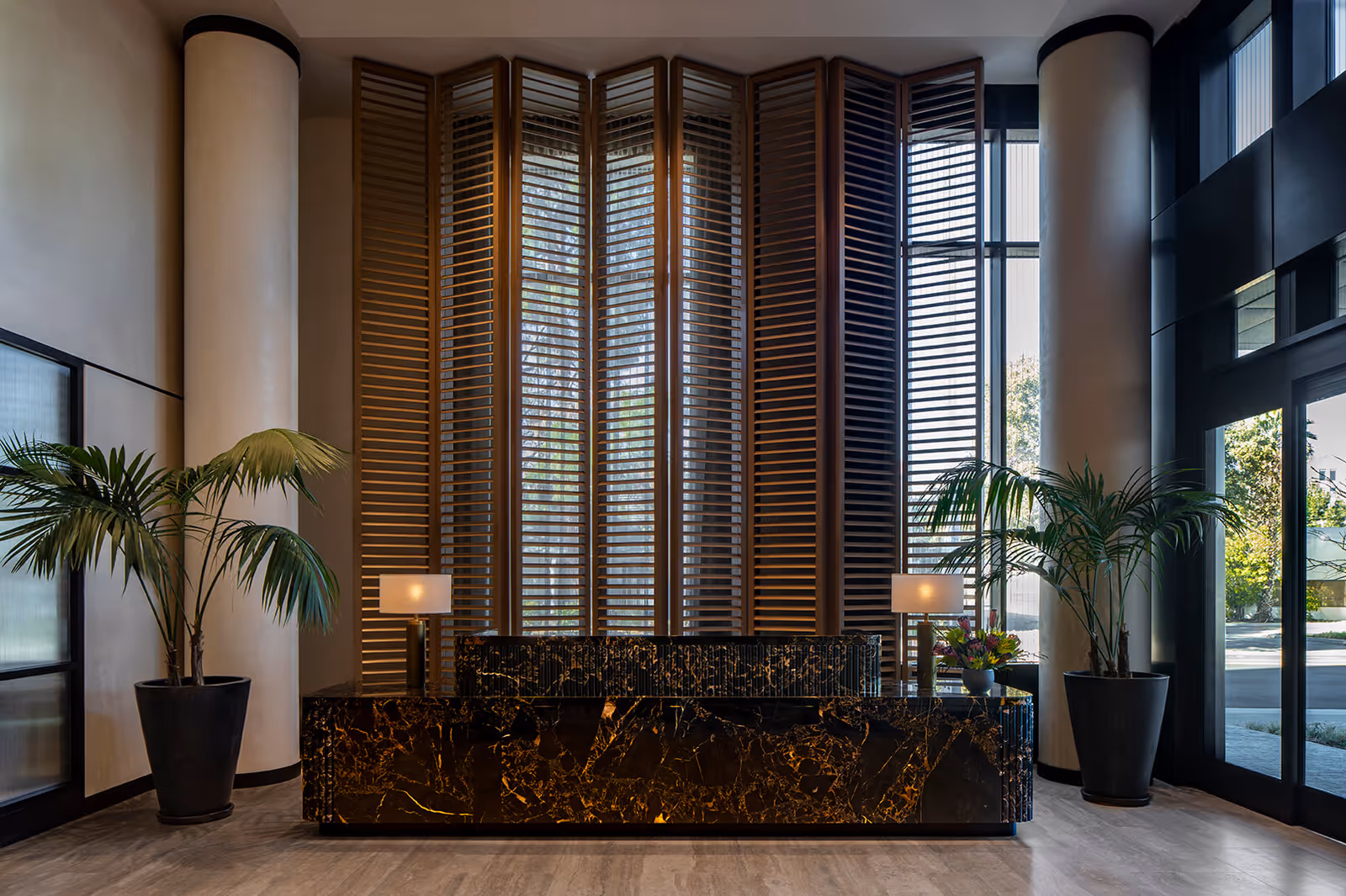 Marble reception desk in a modern lobby with tall wooden shutters, potted palms, and table lamps.