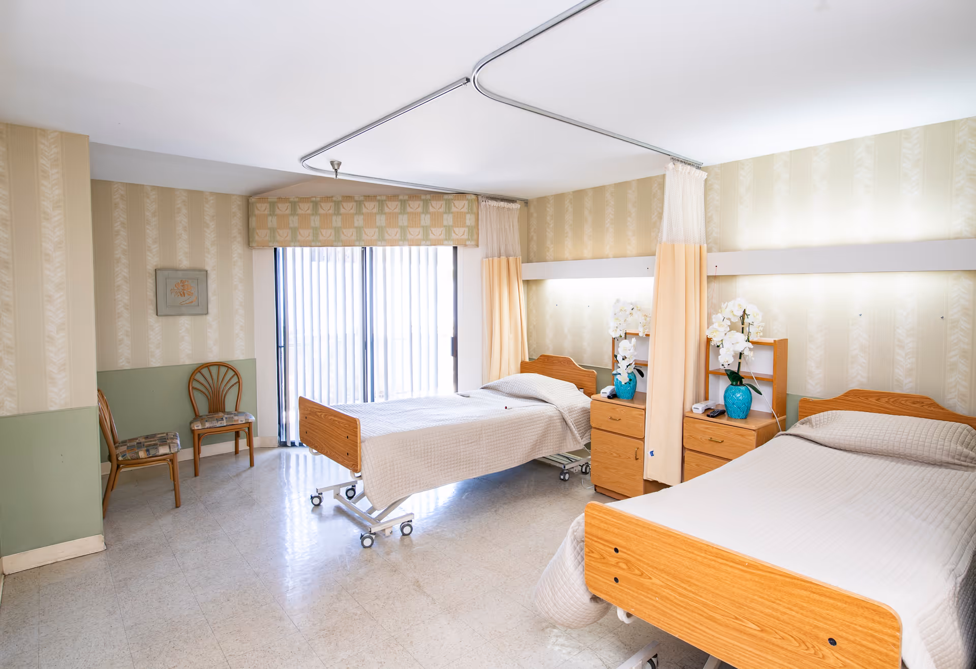 A bright room with two hospital-style beds separated by a privacy curtain. Each bed has a wooden headboard and footboard, beige bedding, and a wooden nightstand with a blue vase holding white flowers. There are two wooden chairs with patterned cushions against the wall, and a large window with vertical blinds letting in natural light. The walls have a beige patterned wallpaper with a green lower section.
