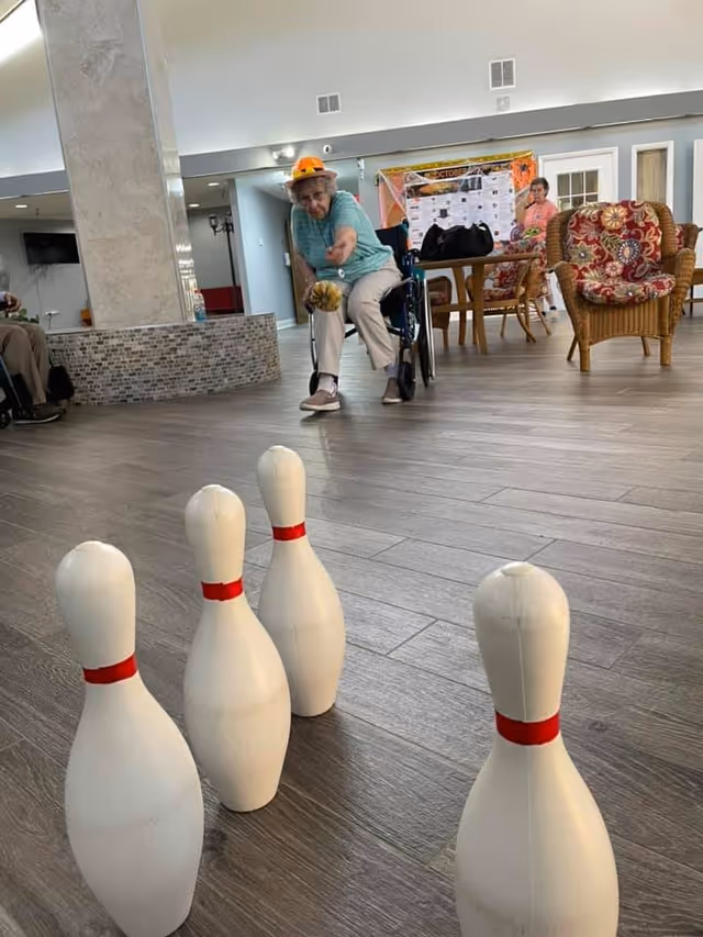 An elderly woman in a wheelchair wearing a hat is rolling a small bowling ball towards five white bowling pins set up on a wooden floor inside a common area. In the background, another elderly woman is seated near a table with chairs and a bulletin board on the wall.