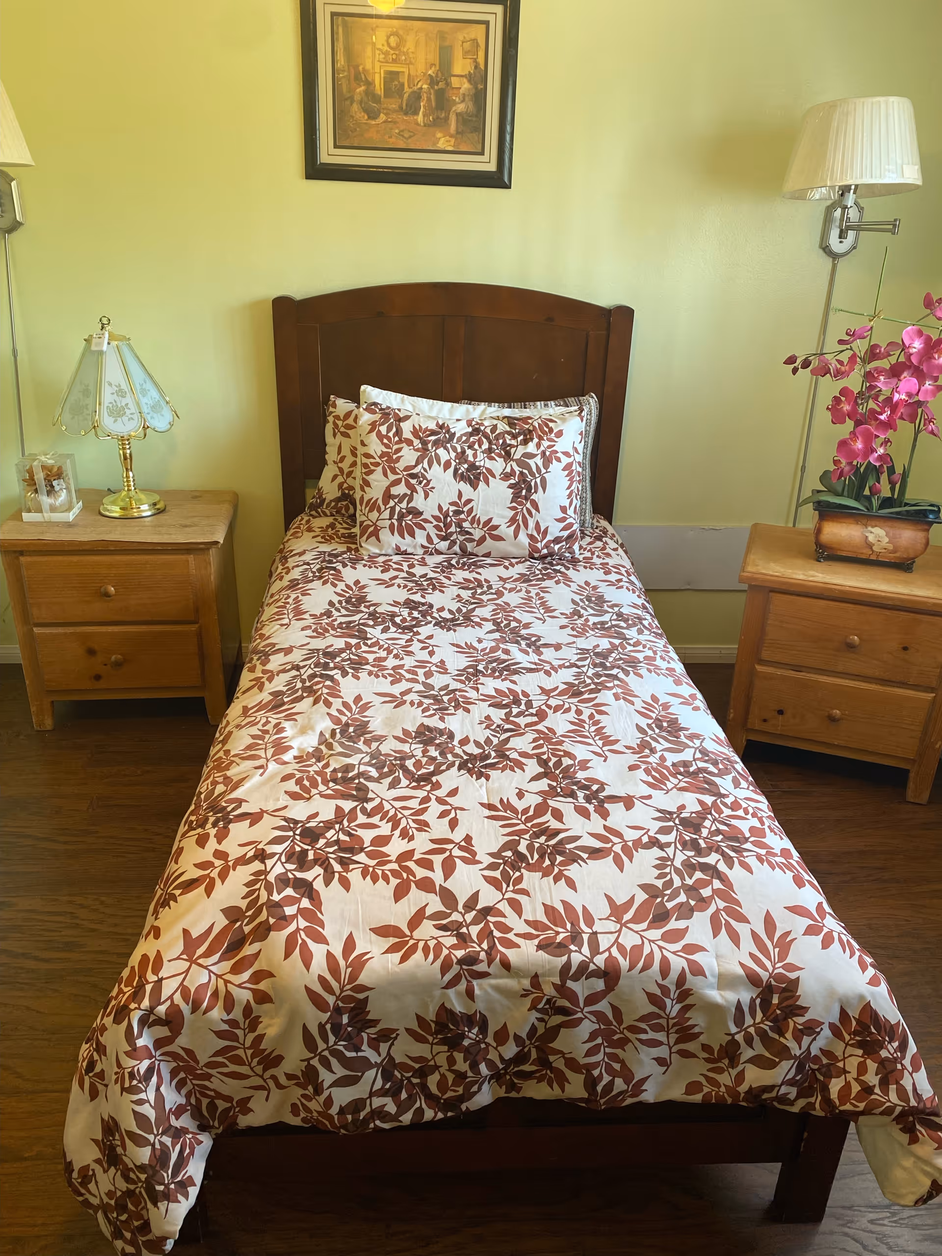 A single bed with a wooden headboard, covered with a white bedspread featuring a brown leaf pattern, and matching pillows. On either side of the bed are wooden nightstands; the left one has a decorative lamp and a small framed photo, while the right one has a potted pink orchid and a wall-mounted lamp above it. A framed painting hangs on the light yellow wall above the bed.