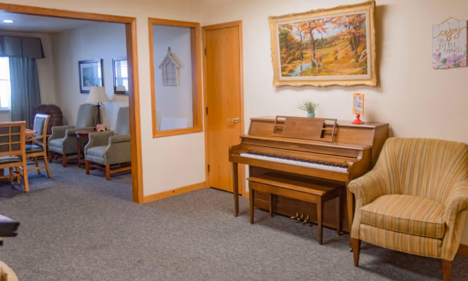 A cozy assisted living common area featuring a wooden upright piano with a bench, a striped armchair, and a framed painting of a deer in an autumn forest hanging above the piano. To the left, there is an open doorway leading to a room with dining chairs, a table, and upholstered armchairs near a window with curtains. The walls are light-colored with wooden trim, and there is a decorative wall hanging that says 'Enjoy the little things.'