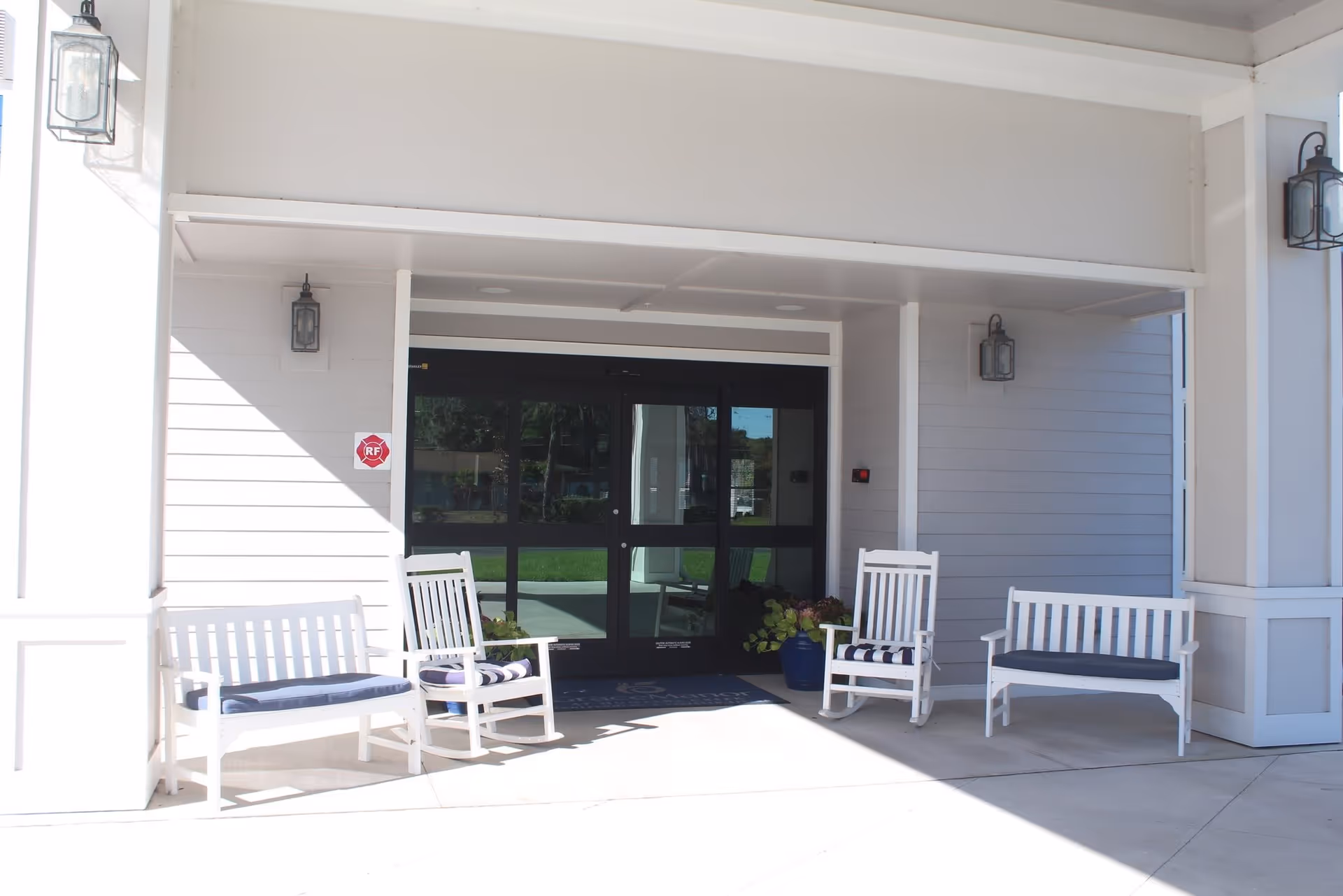 Entrance area of a senior living facility with white benches and rocking chairs with cushions placed on a covered porch. The entrance has double glass doors and is flanked by wall-mounted lantern-style lights. There are potted plants near the doors.