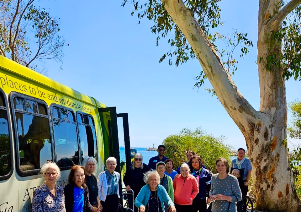 A group of elderly people standing outdoors near a green and yellow bus with trees and a body of water in the background under a clear blue sky.