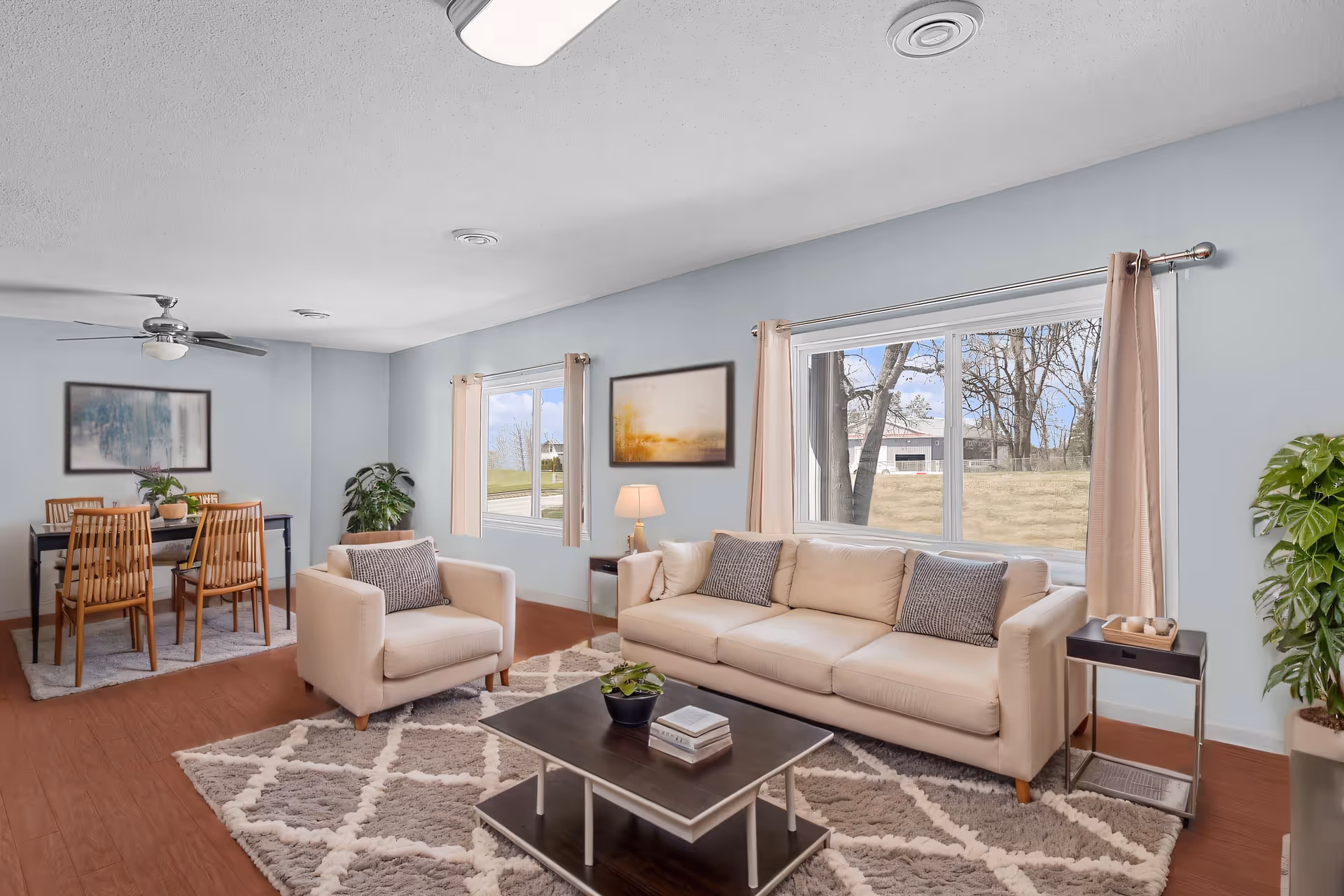 Bright living room with a beige sofa and armchair around a coffee table, a dining table in the background, and large windows showing an outdoor view.