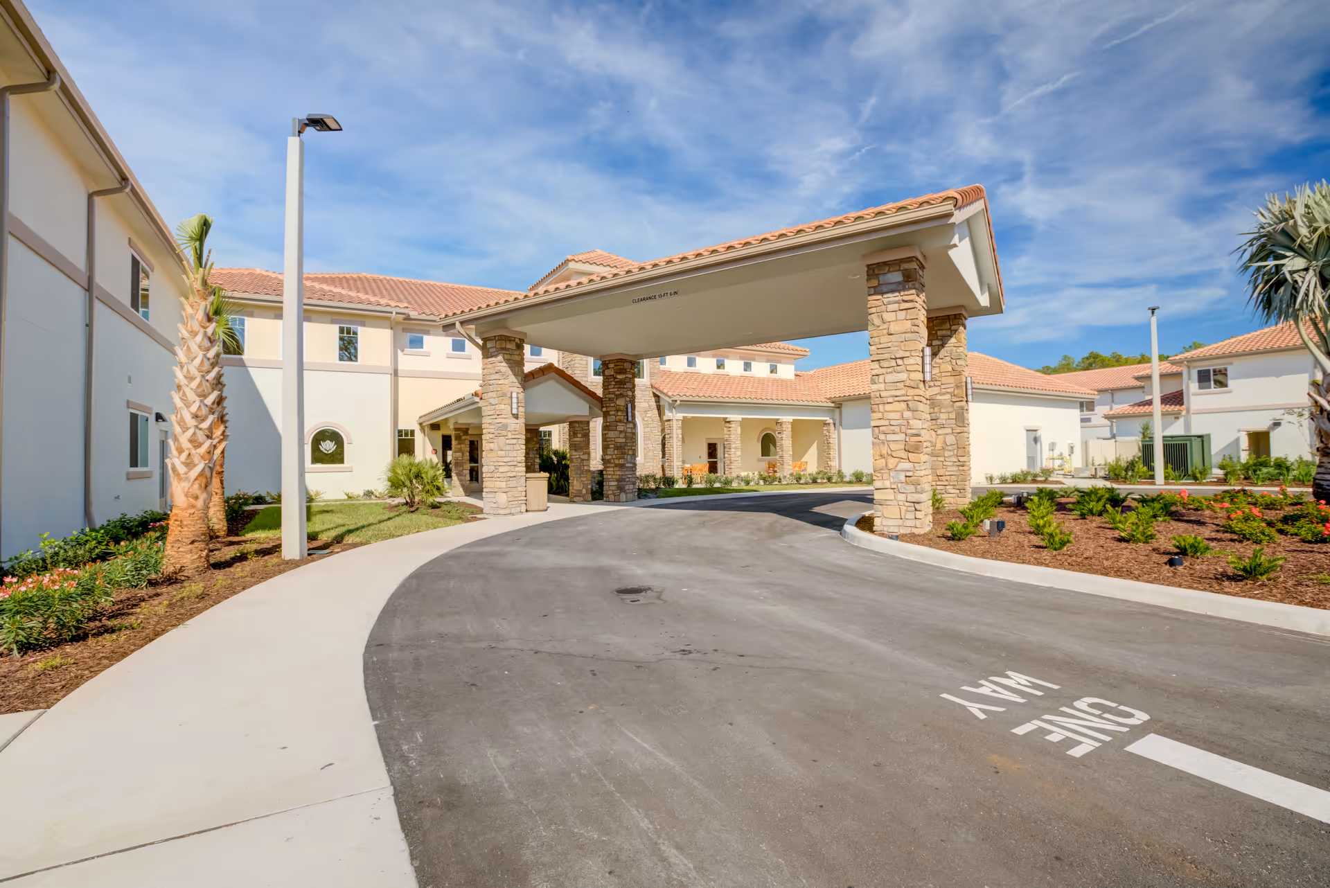 Covered porte-cochère and front entrance of a light-colored building with stone columns, landscaped beds, and a curved driveway.