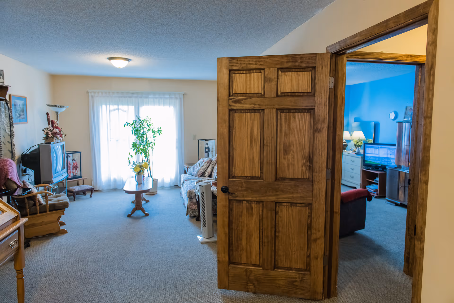 Interior view of a senior living facility showing a living room with a floral couch, wooden coffee table with a flower vase, a TV on a stand, a rocking chair, and a tall plant near a window with sheer curtains. A wooden door is open, revealing a bedroom with blue walls, a TV, and a dresser with lamps and flowers.