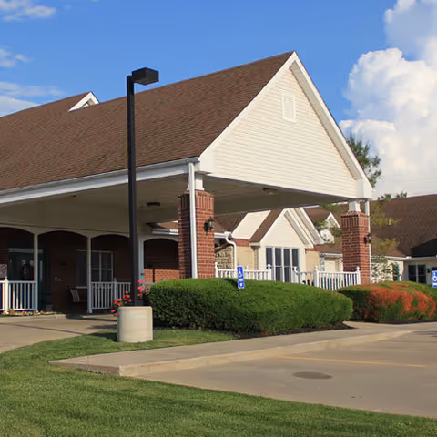 Exterior view of a senior living facility with a covered entrance supported by brick pillars, manicured bushes, and a parking area with handicap parking signs under a partly cloudy sky.