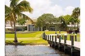 View of a waterfront area with a wooden dock extending over the water, palm trees, green grass, and a building in the background under a partly cloudy sky.